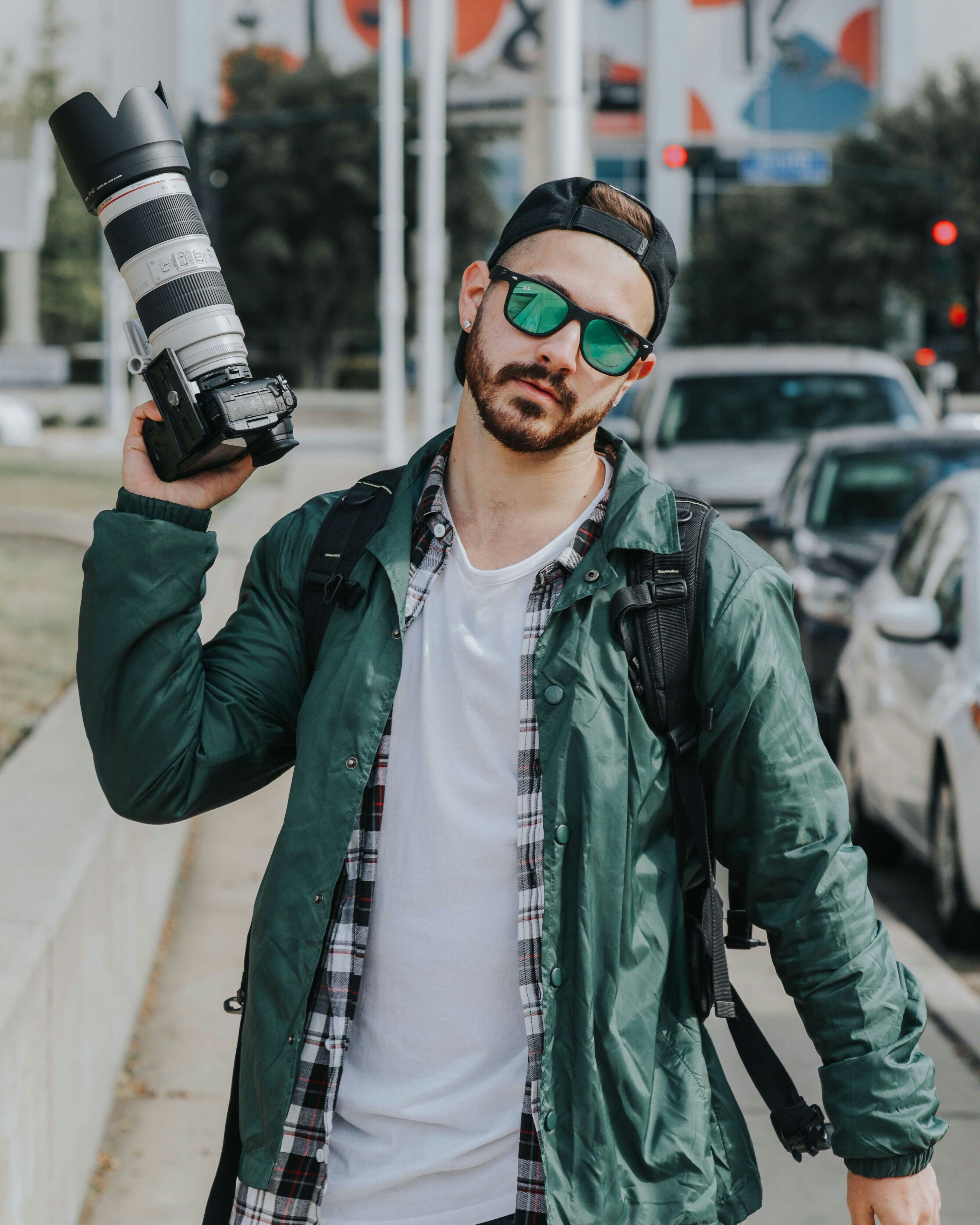 man holding DSLR camera wearing green button-up jacket beside cars